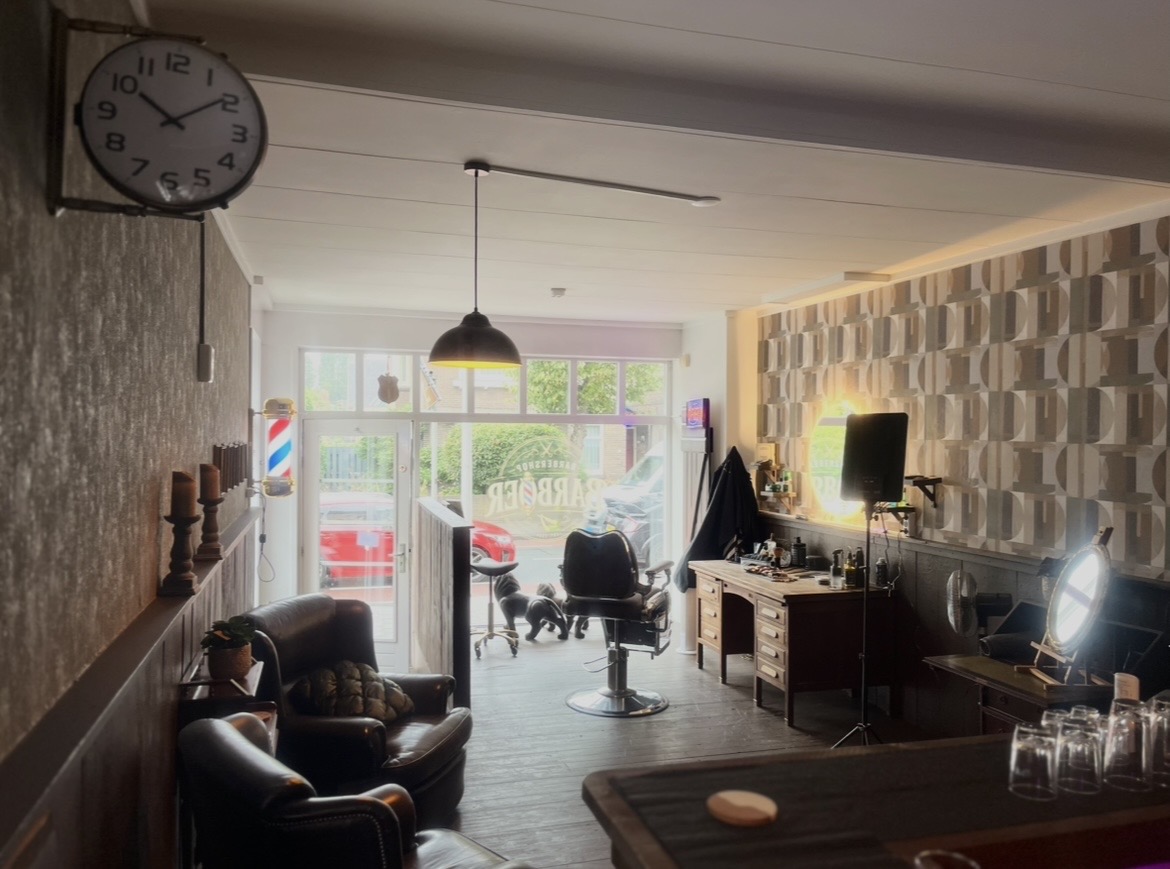 De zaak vanaf de ingang: leren fauteuils, de kappersstoel, een bureau en de klok aan de muur